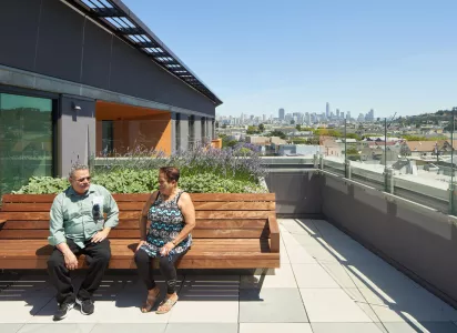 Two people sit on a wooden bench on a rooftop garden, talking under a clear blue sky with a city skyline visible in the background.