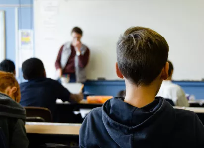 Students sit at their desks facing a teacher at the front of the classroom, with the scene viewed from behind one of the students.