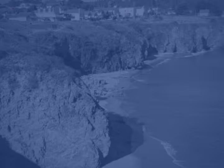 A monochromatic blue aerial view of a coastal cliffside with waves meeting the sandy shore and buildings along the edge above.