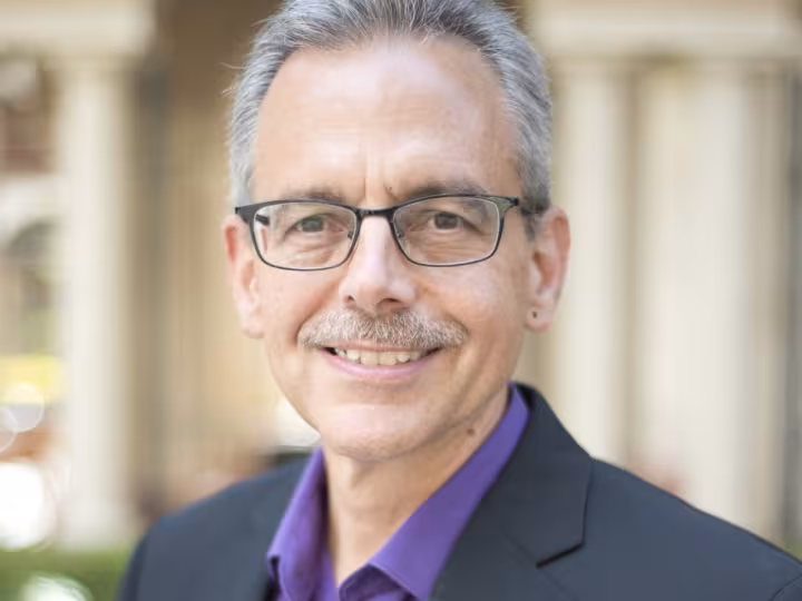 Dr. Manuel Pastor, smiling and wearing glasses, a black blazer, and a purple shirt, standing outdoors in front of an arched building.