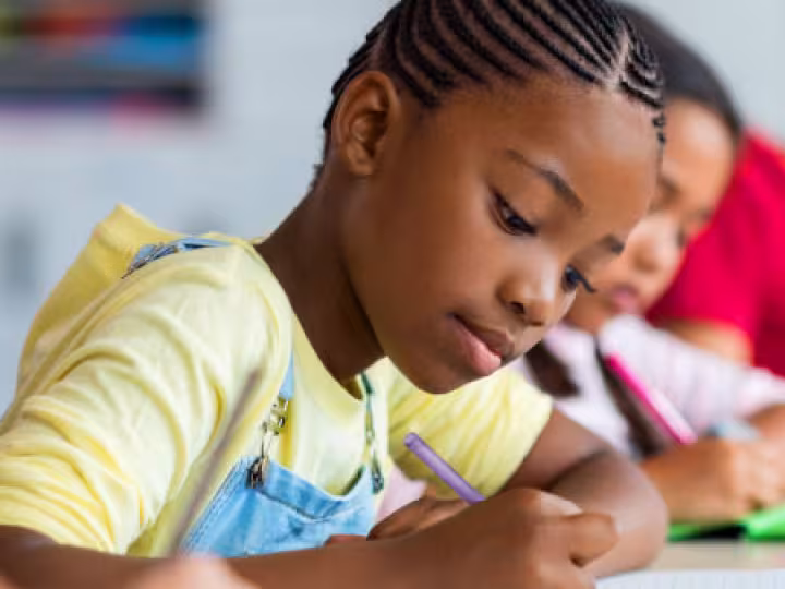 A young girl wearing a yellow shirt writing with a pencil, focusing on her schoolwork.