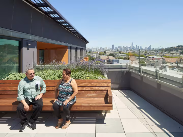Two people sit on a wooden bench on a rooftop garden, talking under a clear blue sky with a city skyline visible in the background.
