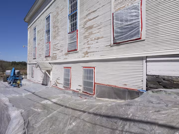 A large white wooden building is shown mid-renovation, with peeling paint, windows sealed in plastic, and protective sheeting covering the ground.